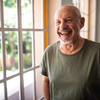 Portrait of casually clothed happy senior man standing near window at home and smilng